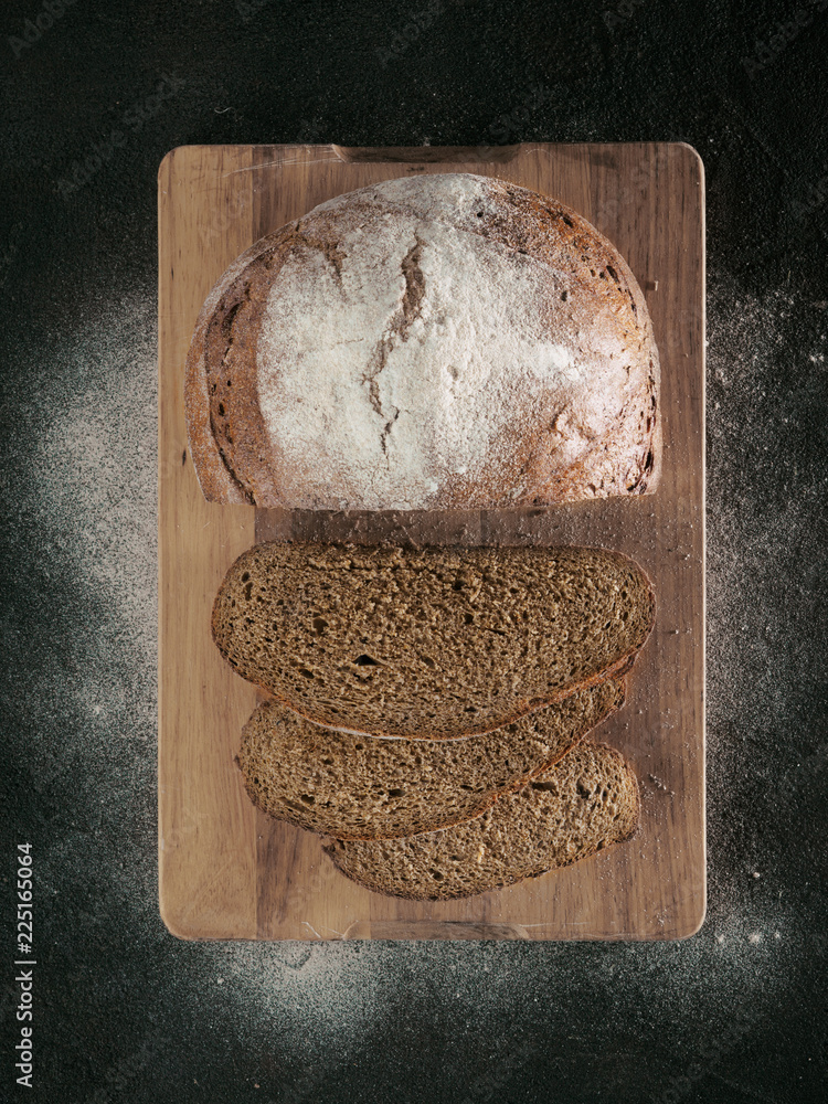 Wall mural sliced homemade sourdough rye bread on cutting board over black textured background with rye flour. 