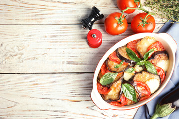 Flat lay composition with baked eggplant, tomatoes and basil in dishware on wooden table. Space for text