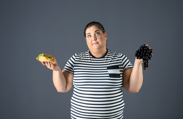 Overweight woman with grapes and hamburger on gray background