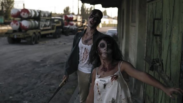 Halloween horror filming concept. Picture of creepy male and female ghost or zombie walking with wounded face and bloody clothes. Industrial, abandoned town and tracks on the background