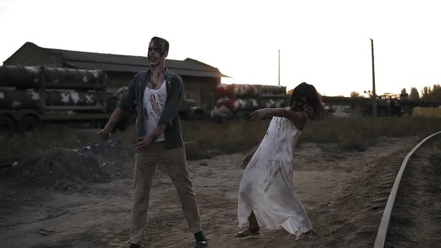 Creepy two zombies in bloody clothes walking through the ruined city during the zombie apocalypse. Abandoned place with trucks with missiles on the background