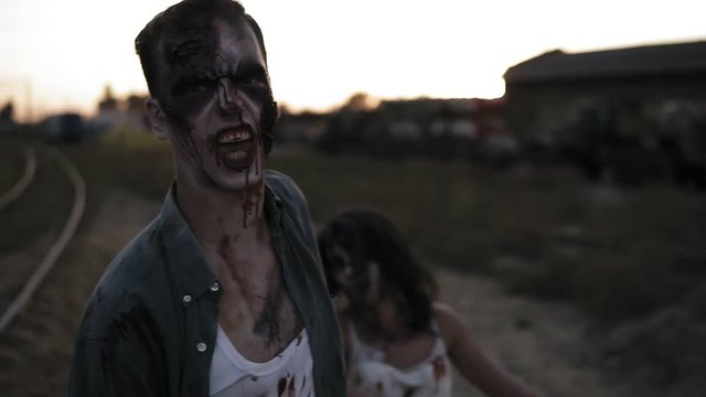 Portrait of a male zombie with bloody teeth and wounded face shouting and zombie girl behind him walking outdoors . Blurred abandoned town on the background