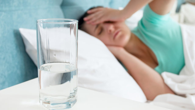 Closeup image of glass of water against woman with headache lying in bed