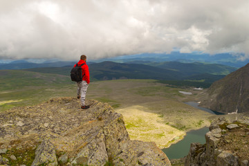 Guy in a red sweatshirt with a backpack is standing with his hands in pockets on the edge of the cliff in front of him is a colorful view of mountains lakes with glaciers. Traveler complete the route