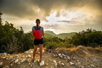 A young girl tourist with a backpack stands on the edge of a mountain and looking into the distance at dawn or sunset