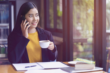 Young attractive business woman talking on smartphone and holding a cup of coffee with happiness and smiling during working at office workplace. Many document or paperwork are on table for analysis