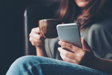 Closeup image of a woman holding , using and touching a smart phone while drinking coffee