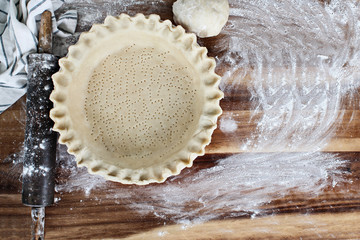 Homemade butter pie crust in pie plate with fluted pinched edge, rolling pin, towel and ball of dough over floured rustic wooden background. Crust has been perforated with fork and ready for baking.