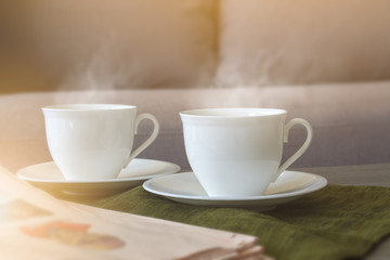 Two white coffee cups with soft smoke stream put on plate and dark green cloth nearly newspaper in the living room and sun lighting from window. Selective focus.