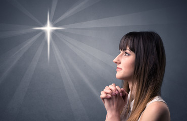 Young woman praying on a grey background with a shiny cross silhouette above her