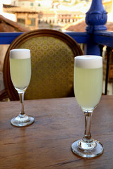 Two glass of Peruvian Pisco Sour on wooden table with a vintage chair and blurred building in background, old town of Cusco, Peru 