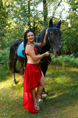 Young beautiful brunette in a red dancer costume next to a black horse in the forest. Overall plan.