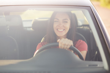 Satisfied attractive woman learns drive car, being in good mood as has positive resuls, drives by herself, keeps hands on wheel, has pleasant smile, dressed in casual clothes. Delighted driver