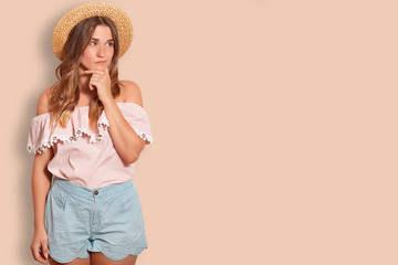 Thoughtful European female traveller holds chin and looks pensively aside, dressed in summer straw hat, blouse and shorts, poses against beige background with copy space for your advertisement