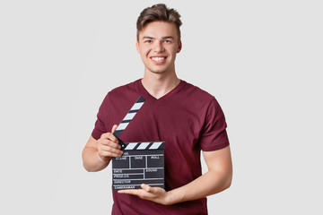 Photo of attractive guy with stylish hairdo, holds clapperboard, being producer of new film, smiles...
