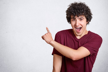 Positive curly guy keeps mouth opened, exclaims with happiness, points aside at blank copy space...