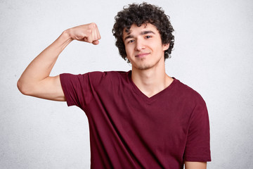 Satisfied strong man shows biceps or muscles, has delighted expression, curly hairstyle, trains every day to achieve aims and have sporty body, poses against white background, feels proud of himeself