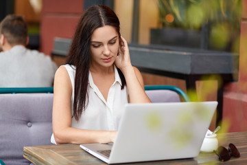 Photo fo concetrated female freelancer watches tutorial online, uses laptop computer for work, sits at table in outdoor cafeteria, waits for her order. Woman makes shopping on website with discounts