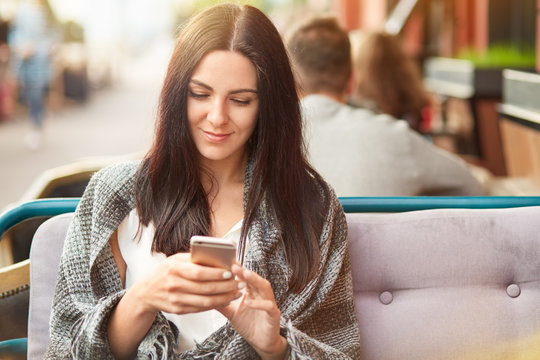 Online internet connection concept. Beautiful young brunette lady uses mobile phone for searching internet, wrapped in coverlet, spends free time in coffee shop, recieves message from boyfriend