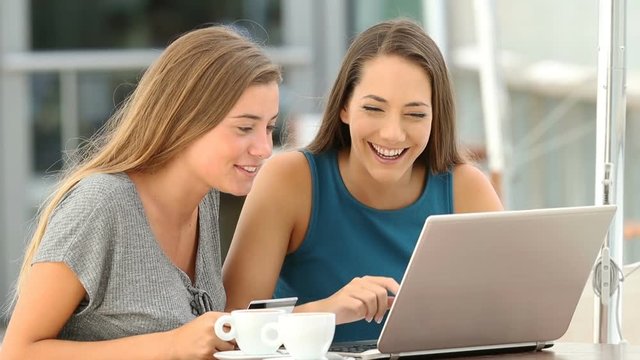 Two happy friends shopping online with a laptop paying with a credit card in a coffee shop