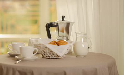 Coffee maker, cups and croissants in the basket are on a round table in front of the window.
