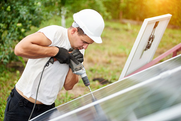 Profile of professional technician working with screwdriver connecting solar photo voltaic panel to metal platform on green summer blurred bokeh background. Alternative sun energy production concept.