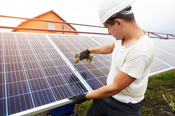 Professional technician working with screwdriver connecting solar photo voltaic panel to exterior metal platform under clear blue sky. Alternative renewable ecological green energy concept.
