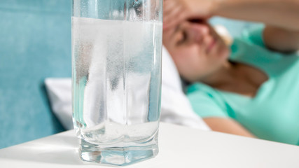 Closeup image of air bubbles in glass of water with aspirin on bedside table next to woman suffering from headache in bed