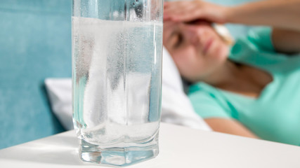 Closeup image of glass of carbonated water on bedside table next to sick woman lying in bed