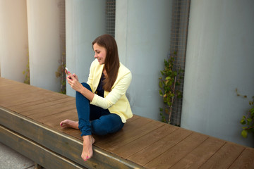 beautiful young girl sitting on a bench in a yellow jacket with a phone in her hand in summer