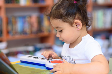 Little Child Indoors In Front Of Books. Cute Young Toddler Sitting On A Chair Near Table and play with toy piano. Kid in a bookstore, surrounded by colorful books. Happy multirace girl play the piano.