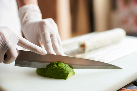 Close-up view of process of preparing delicious rolling sushi in restaurant. Female hands in disposable gloves slicing avocado on wooden board with the knife.