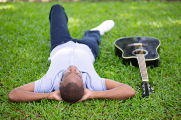Black man lying upside down on grass and guitar. Handsome young man in park. Guitarist and rest...