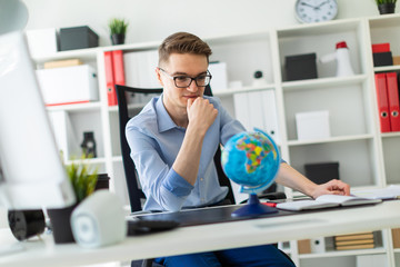 A young man sits in the office at a computer desk and in front of him stands a globe.