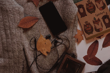 Fototapeta premium Perfume, mobile phone, headphones and yellow leaves on the cozy autumn sweaters top view.