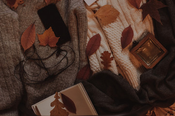 Fototapeta premium Autumn cozy flatlay. Smartphone with headphones, whiite ntebook and perfume on the autumn sweaters with scarf
