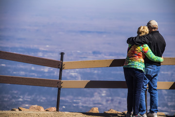 An older couple stand with their arms around each other at the sumit of a mountains looking down at...
