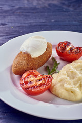 A full-fledged hot dinner on a white plate in a cafe, restaurant. Chicken Kiev stuffed with breaded, poached eggs, grilled tomato, arugula and mashed potatoes for dinner.