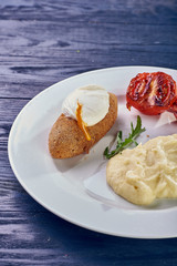 A full-fledged hot dinner on a white plate in a cafe, restaurant. Chicken Kiev stuffed with breaded, poached eggs, grilled tomato, arugula and mashed potatoes for dinner.