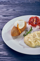 A full-fledged hot dinner on a white plate in a cafe, restaurant. Chicken Kiev stuffed with breaded, poached eggs, grilled tomato, arugula and mashed potatoes for dinner.