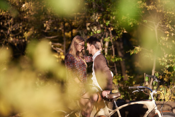Fototapeta na wymiar Smiling romantic couple, bearded man and attractive woman close together at tandem double bicycle outdoors in dark autumn park or forest on sunny clearing on blurred golden green bokeh background.