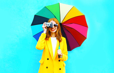 Happy smiling woman takes a picture on retro film camera holds colorful umbrella in yellow coat...