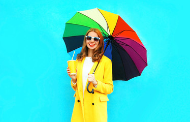 Happy smiling woman with cup of fruit juice, colorful umbrella in yellow coat jacket on blue background
