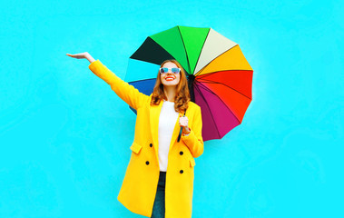 Happy smiling woman with colorful umbrella in yellow coat enjoys on blue background