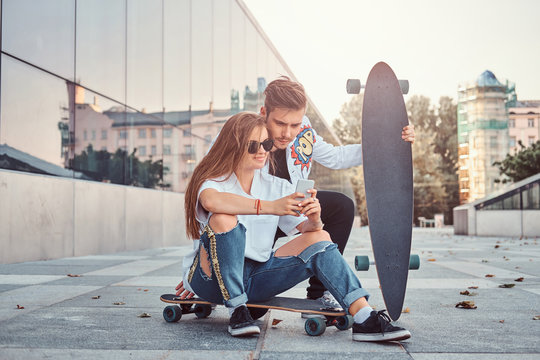 Trendy dressed couple with skateboards views interesting photos together on smartphone on the street.