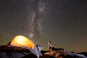 Athletic girl doing push-up exercises on big boulder on night starry sky with Milky way and brightly lit tourist tent background. Active lifestyle and mountain camping concept. Urdhva Mukha Shvanasana