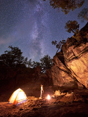 Campsite at night amid huge rock formations. Athletic slim barefooted girl standing straight between small tourist tent and burning bonfire under clear dark starry sky. Tourism and camping concept.