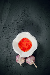 Top view of a white bowl with red sauce and garlic next to it. Red sauce and garlic on a stone, dark table. The concept of preparing dishes, illustrative photo for food photography.