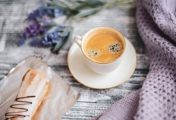 Cup of hot coffee, eclair and flowers on rustic wooden table, closeup photo warm sweater, winter morning concept, top view. Vintage tinted