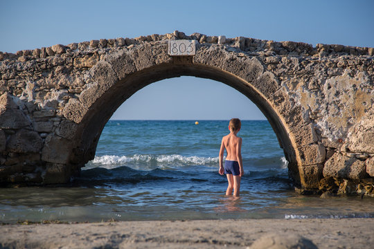Young boy in swimsuit stands under an old stone bridge in water of sea, in Greece 